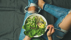  Green vegan breakfast meal in bowl with spinach, arugula, avocado, seeds and sprouts. Girl in jeans holding fork with knees and hands visible, top view, copy space. Clean eating, vegan food concept (Green vegan breakfast meal in bowl with spinach, aru 